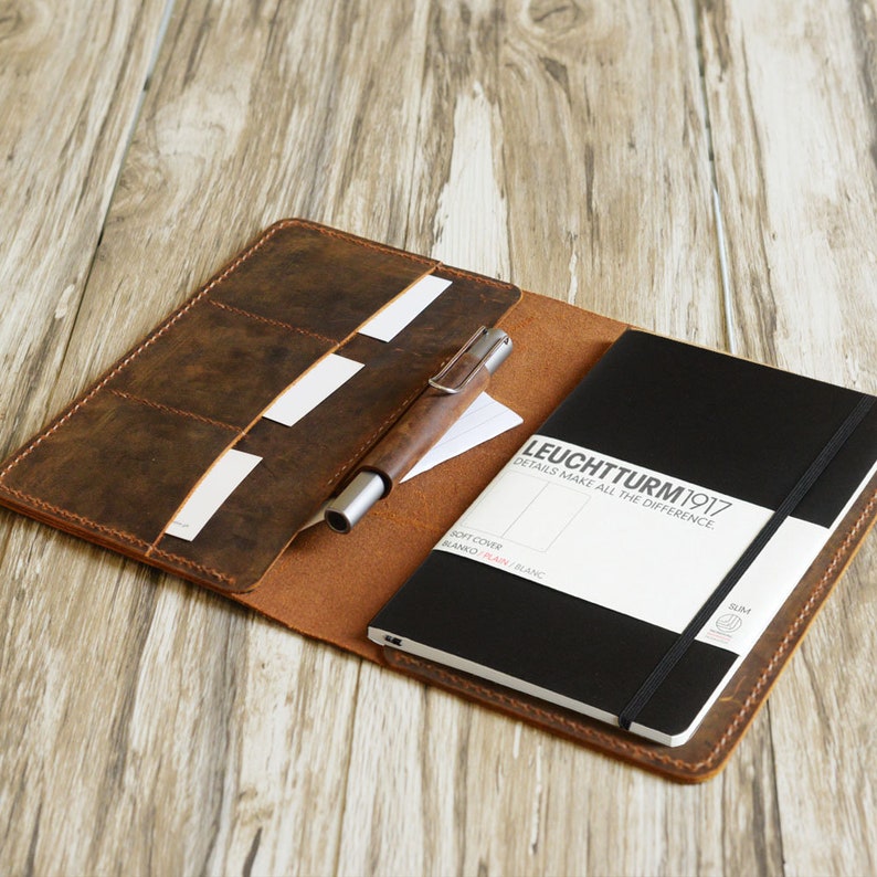 May include: A brown leather notebook cover with a black notebook inside. The notebook has the text "LEUCHTTURM1917" and "DETAILS MAKE ALL THE DIFFERENCE." The cover has multiple slots for cards and a pen holder.