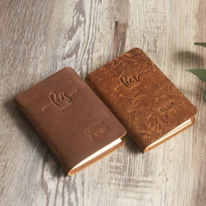 May include: Two brown leather-bound journals with embossed floral designs. One journal says "his vows" and "DAMEN 8.11.22". The other journal says "her vows" and "EMMA 8.11.22".
