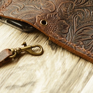 K&ouml;nnte beinhalten: Eine braune Leder Handyh&uuml;lle mit einem gepr&auml;gten Blumenmuster. Die H&uuml;lle hat einen Metallverschluss und ein Loch f&uuml;r einen Riemen.