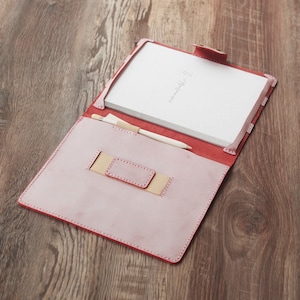 Puede incluir: Una cubierta de cuaderno de cuero rosa con un portaplumas y un cuaderno blanco en el interior. El cuaderno tiene la palabra "remarkable" escrita en él.