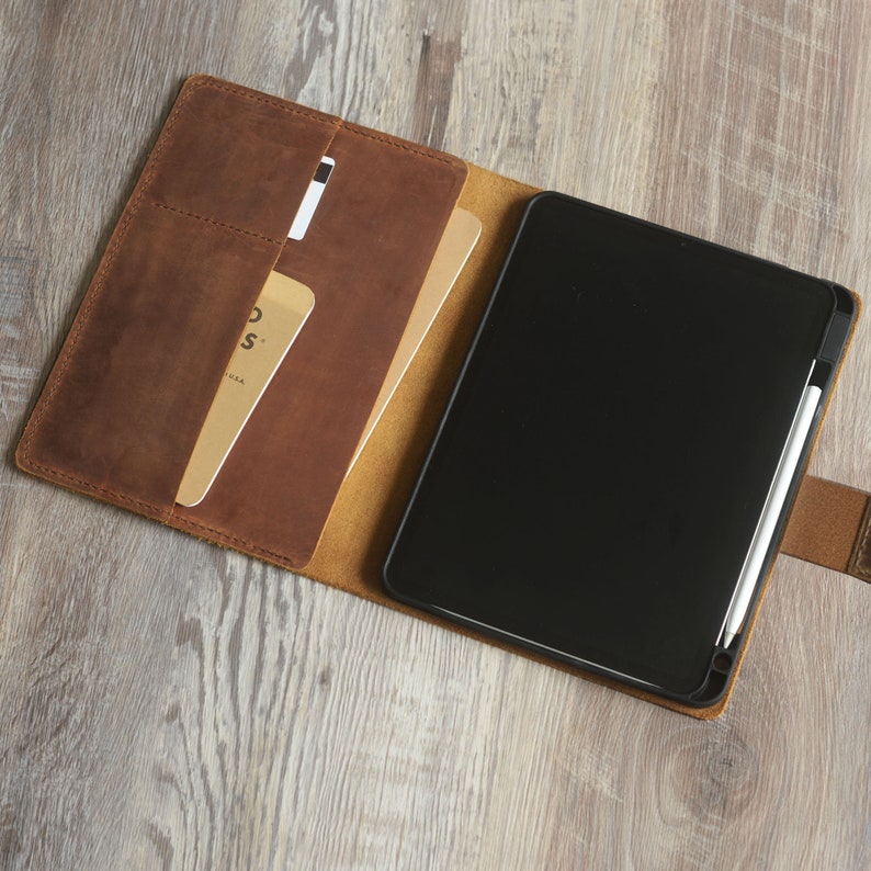 以下が含まれることがあります： 黒いタブレットと白いスタイラスが入った茶色の革製フォリオケース。ケースにはカードやノート用のポケットが複数あります。