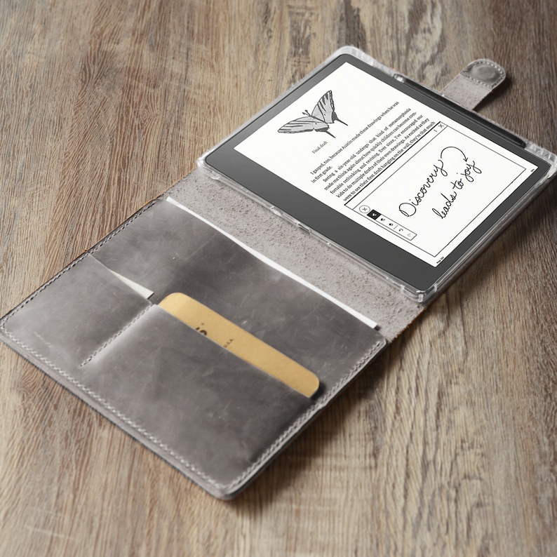 Op de afbeelding: Een grijs leren etui met een ingebouwd vak voor een stylus. De etui is open en een zwarte e-reader bevindt zich erin. Het scherm van de e-reader toont een vlindergrafiek en de tekst "Discovery leads to joy".