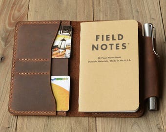 May include: A brown leather notebook cover with a pen holder and two card slots. The cover is open to reveal a Field Notes memo book with the text "48-Page Memo Book Durable Materials/Made in the U.S.A."