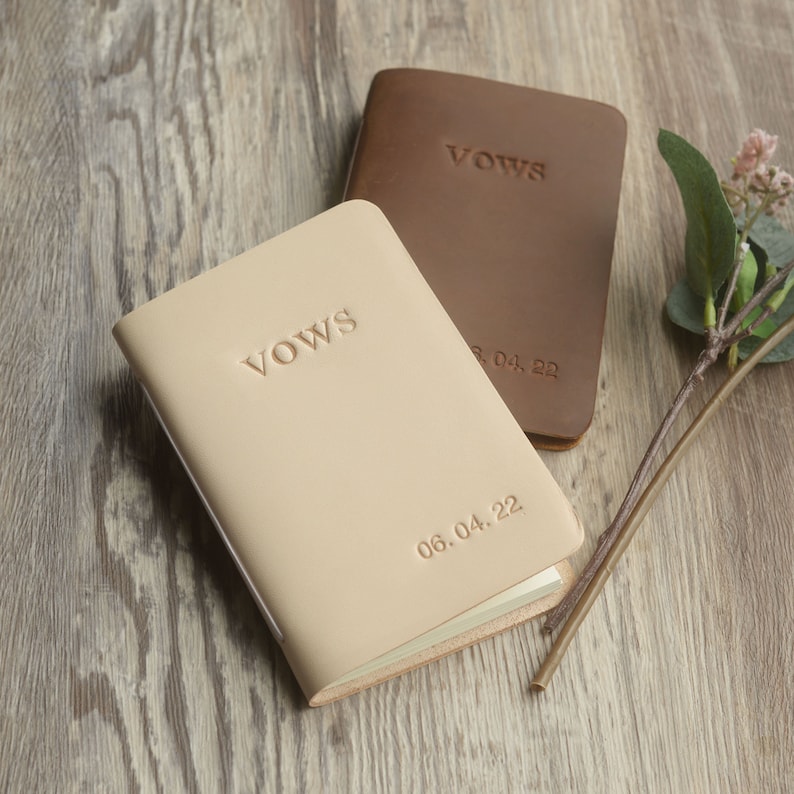 May include: Two leather-bound vow books, one beige and one brown, with the word "Vows" embossed on the front. The beige book has the date "06.04.22" embossed on the front. The brown book has the date "28.04.22" embossed on the front.