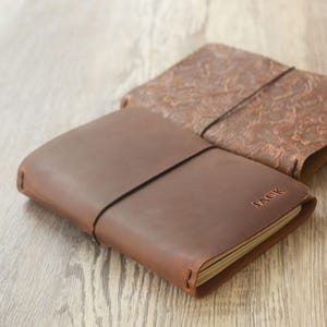 Op de afbeelding: Twee bruine leren notitieboeken. Het voorste notitieboek heeft een donkerdere bruine tint met de naam "JACK" in reliëf op de kaft. Een zwarte elastische band loopt om het notitieboek. Het tweede notitieboek is lichtbruin met een bloemreliëf.