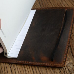 May include: A brown leather notebook cover with a black elastic closure. The cover has a distressed look and is open to reveal lined pages.