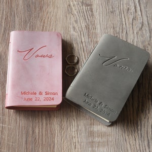 Peut inclure: Deux livres de v&oelig;ux reli&eacute;s en cuir avec le mot &laquo; V&oelig;ux &raquo; en relief sur la couverture. Les livres sont rose et gris et portent les noms Michele & Simon ainsi que la date du 22 juin 2024.