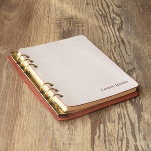 Op de afbeelding: Een bruin lederen gebonden notitieboek met gouden ringen en een witte pagina met de tekst "Lorem Ipsum" erop gedrukt.