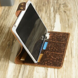 May include: A brown leather tablet case with a floral embossed design. The case has a stand feature and slots for cards and earphones. A tablet is shown in the case.
