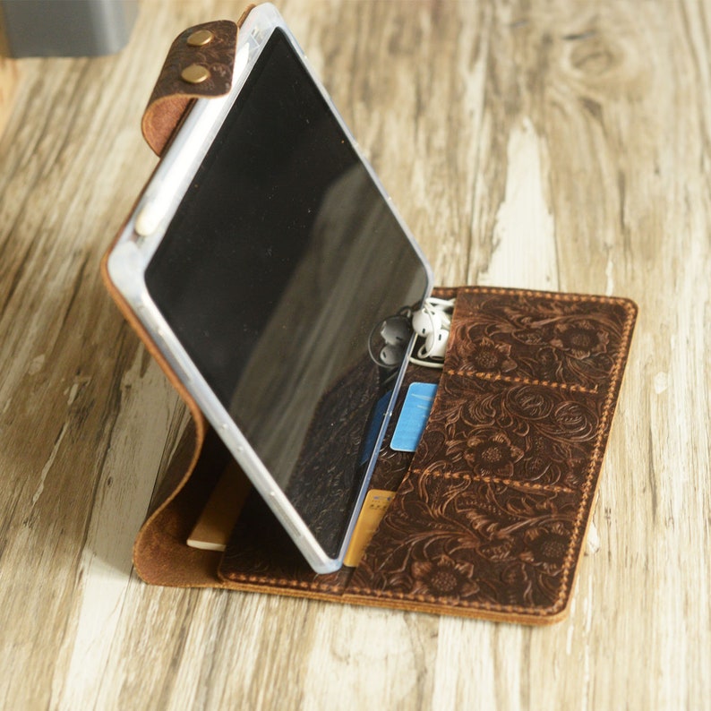 May include: A brown leather tablet case with a floral embossed design. The case has a stand feature and slots for cards and earphones. A tablet is shown in the case.