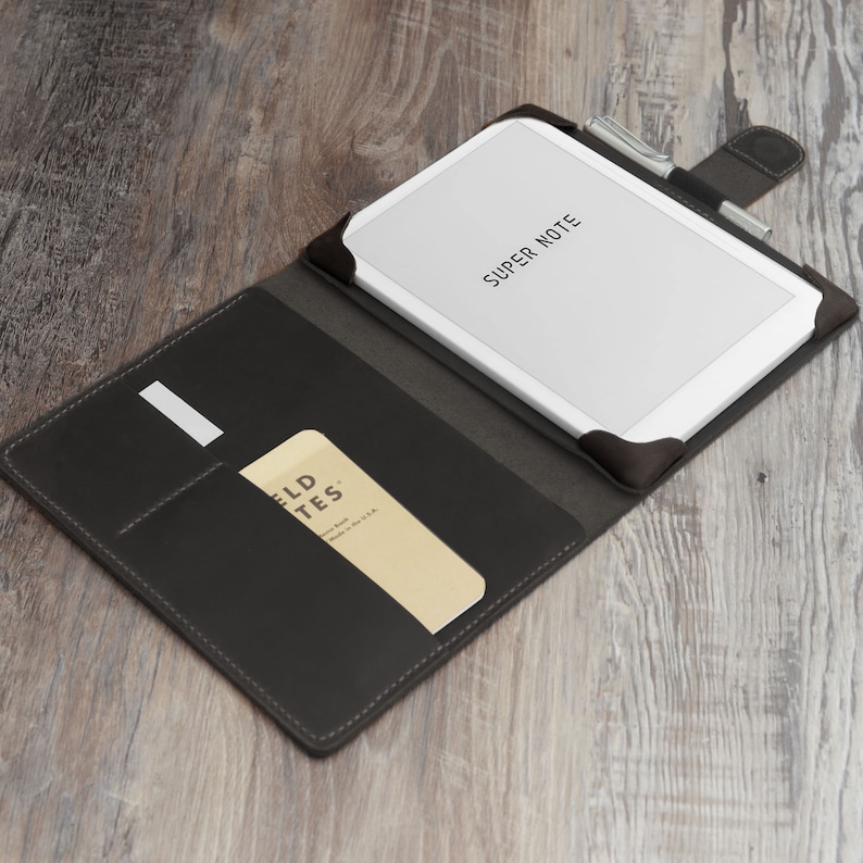 May include: A black leather notebook cover with a white tablet inside, featuring the text "SUPER NOTE." A pen is secured at the top. A small notepad is in the pocket, with the text "FIELD NOTES."