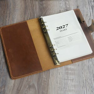 Può includere: Una copertina per diario in pelle marrone con un meccanismo di rilegatura in metallo a sei anelli che trattiene pagine bianche. La pagina superiore presenta il testo 2027 DIARY e una sezione per le informazioni personali.