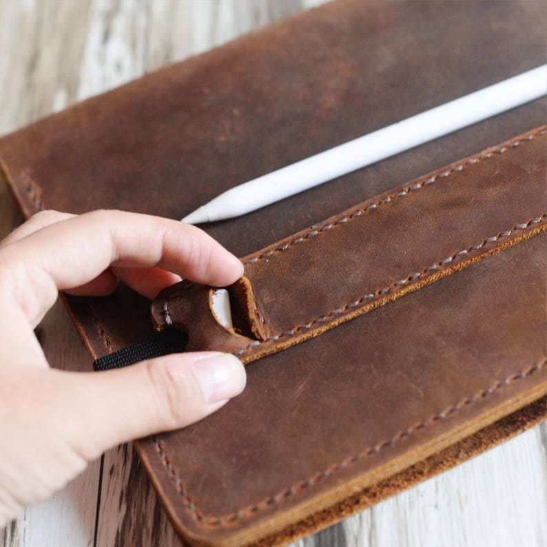 May include: A brown leather notebook cover with a white stylus tucked into a leather strap. The cover has stitching around the edges and a flap that closes with a snap.