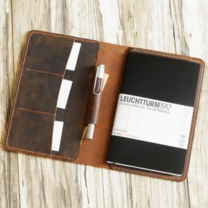 May include: A brown leather notebook cover with a pen and a black notebook inside. The notebook has the text "LEUCHTTURM1917" and "DETAILS MAKE ALL THE DIFFERENCE." on the front cover.