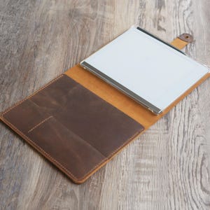 May include: A brown leather portfolio with a clear plastic insert for a tablet or notepad. The portfolio has a flap that closes with a snap and features a pocket on the front.