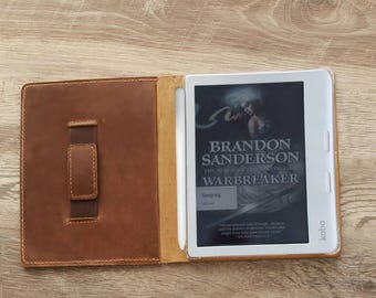 Puede incluir: Una funda de cuero marrón para lector electrónico con correa de mano y soporte para lápiz óptico blanco, que contiene un lector electrónico Kobo blanco con la portada del libro Warbreaker de Brandon Sanderson. La funda está abierta.