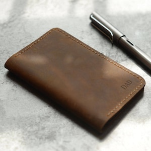 May include: A brown leather notebook cover with the word "DAD" embossed on the front. The cover is lying on a gray surface next to a silver pen.