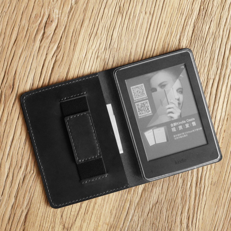 May include: Black leather case with a built-in card slot for a Kindle Oasis e-reader. The case is open and the e-reader is visible with a screen displaying a promotional image for the Kindle Oasis.