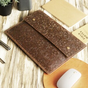 Peut inclure: Une pochette pour ordinateur portable en cuir marron avec un motif en relief floral. La pochette a une fermeture &agrave; rabat avec un bouton en laiton. La pochette est sur une surface en bois avec une souris et un carnet.