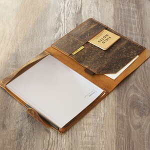 Puede incluir: Un portafolio de cuero marr&oacute;n con un dise&ntilde;o estampado floral. El portafolio tiene un bolsillo para una libreta y un portaplumas. La libreta est&aacute; en blanco y la pluma es negra. El texto "remarkable" est&aacute; impreso en la libreta.
