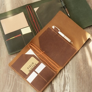 Peut inclure: Deux protège-carnets en cuir, l'un vert foncé et l'autre beige, dotés de poches intérieures, d'élastiques pour maintenir les inserts et de passants pour stylo. La couverture beige contient un carnet Field Notes, avec des stylos et des fentes.