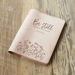 Może przedstawiać: Różowy skórzany notes z wygrawerowanymi słowami "Be still and know that I am God. Psalm 46:10" oraz imieniem "Marie Magee" na okładce. Notes ozdobiony jest wzorem kwiatowym.