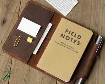 May include: A brown leather notebook cover with a pen holder and a Field Notes memo book inside. The cover has a pocket for cards and a small yellow sticky note.
