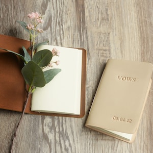 May include: Two leather-bound journals, one brown and one beige. The beige journal has the word "VOWS" embossed on the front cover and the date "06.04.22" printed below.