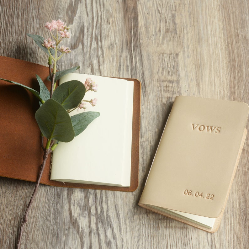 May include: Two leather-bound journals, one brown and one beige. The beige journal has the word "VOWS" embossed on the front cover and the date "06.04.22" printed below.