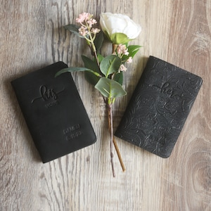 May include: Two black leather vow books with embossed floral designs. The book on the left says "his vows" and the book on the right says "her vows".