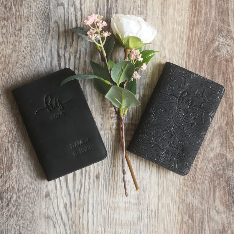 May include: Two black leather vow books with embossed floral designs. The book on the left says "his vows" and the book on the right says "her vows".