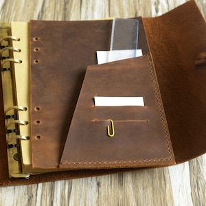 May include: A brown leather bound journal with a ring binder and a pocket on the inside cover. The journal is open to reveal a blank page and a clear plastic ruler.