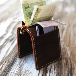 May include: A brown leather wallet with a flap that opens to reveal a cash pocket and card slots. The wallet is sitting on a wooden surface with some bills sticking out of the cash pocket.