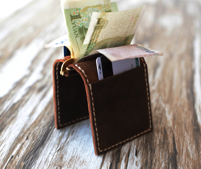 May include: A brown leather wallet with a flap that opens to reveal a cash pocket and card slots. The wallet is sitting on a wooden surface with some bills sticking out of the cash pocket.