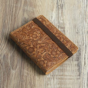 Peut inclure: Un carnet en cuir marron avec un motif en relief floral et une fermeture &eacute;lastique marron.