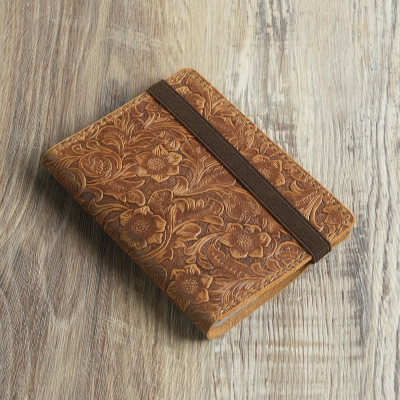 Peut inclure: Un carnet en cuir marron avec un motif en relief floral et une fermeture &eacute;lastique marron.