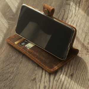 May include: A brown leather phone case with an embossed floral design. The case has a slot for a credit card and a flap that folds over the phone.