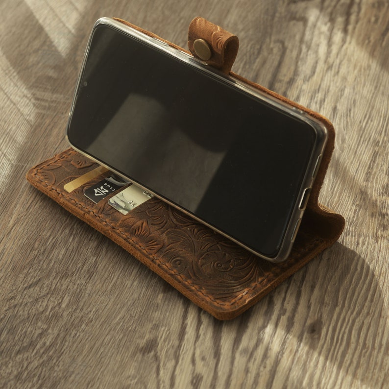 May include: A brown leather phone case with an embossed floral design. The case has a slot for a credit card and a flap that folds over the phone.