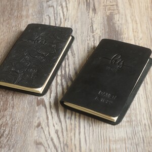 May include: Two black leather journals with embossed text. One journal says "her vows" with floral designs and the name "Emma" and the date "8.11.23". The other journal says "his vows" with the name "Damen" and the date "8.11.23".
