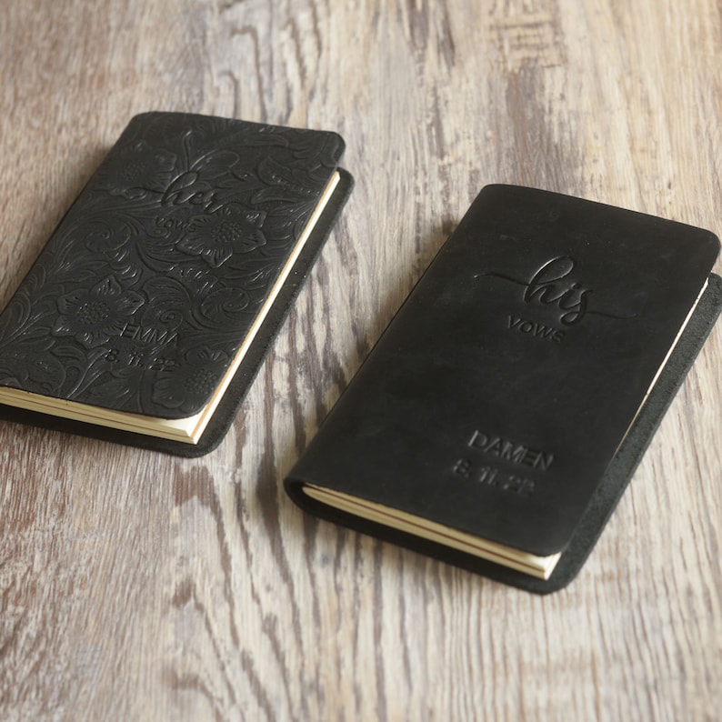May include: Two black leather journals with embossed text. One journal says "her vows" with floral designs and the name "Emma" and the date "8.11.23". The other journal says "his vows" with the name "Damen" and the date "8.11.23".