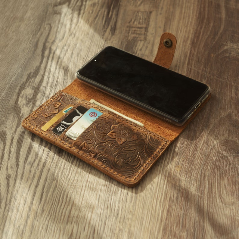 May include: A brown leather phone case with a floral embossed design. The case is open and has a smartphone and credit cards inside.