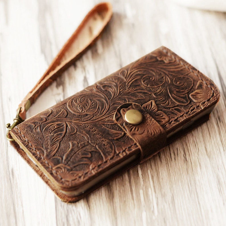 K&ouml;nnte beinhalten: Eine braune Leder-Handyh&uuml;lle mit einem gepr&auml;gten Blumenmuster und einem Riemen. Die H&uuml;lle hat einen Knopfverschluss aus Messing.