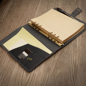 May include: A black leather bound journal with a gold ring binder and a yellow notepad inside. The journal has a pocket on the inside cover and a gold paperclip.