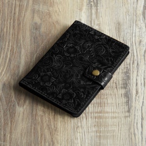 Peut inclure: Un portefeuille en cuir noir avec un motif en relief floral et une fermeture &agrave; bouton dor&eacute;.