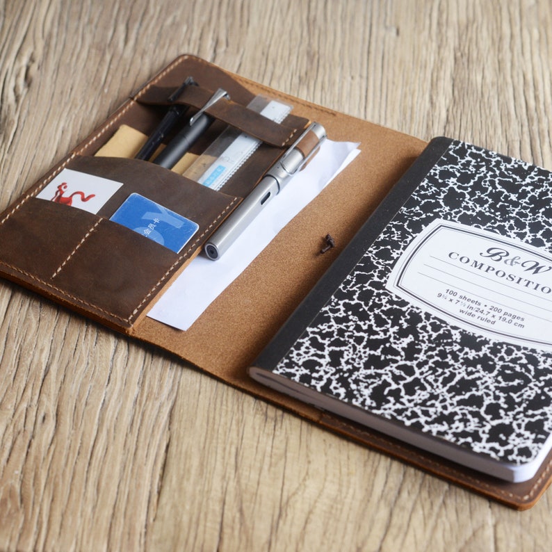 May include: A brown leather notebook cover with a pen, ruler, and cards inside. The cover has multiple pockets and slots for organisation. The notebook is black with white speckled paper and says "B&W COMPOSITIO 100 sheets - 200 pages 9 x 7 in/24.7 x 19.0 cm wide ruled".