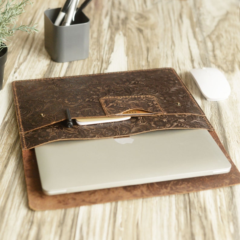 Peut inclure: Une pochette pour ordinateur portable en cuir marron avec un motif floral en relief. La pochette est ouverte et un ordinateur portable est &agrave; l'int&eacute;rieur. La pochette a une poche sur le devant.