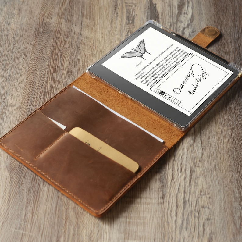 Op de afbeelding: Een bruine leren folio-hoes met een ingebouwd vak voor een kaart of notitieblok. De hoes is open en een zwarte e-reader bevindt zich erin. Het scherm van de e-reader toont een vlinderbeeld en de tekst "Discovery leads to joy."
