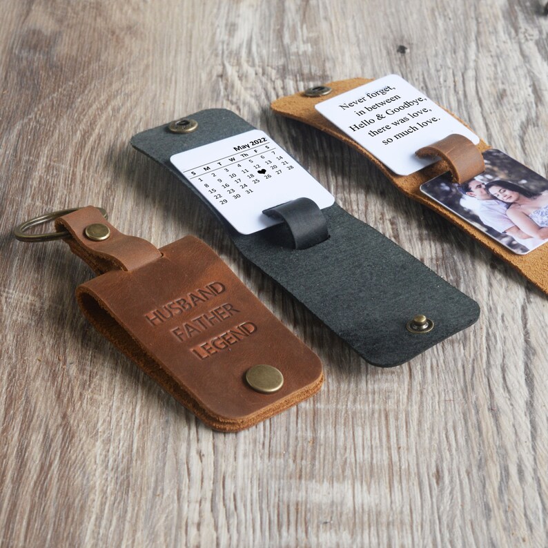 May include: Three leather keychains with different designs. The first keychain is brown and has the words "Husband Father Legend" embossed on it. The second keychain is black and has a small calendar card with the month of May 2022. The third keychain is brown and has a small photo card.