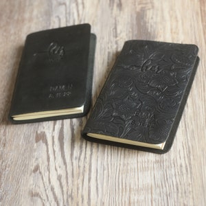 May include: Two black leather journals with personalized engravings. One journal has a floral design and the text "her" and "Emma 8.11.22". The other journal has the text "our love" and "Damen 8.11.22".