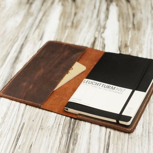 May include: A brown leather notebook cover with a black notebook inside. The notebook has a black elastic band and a white label that says "LEUCHTTURM1917 DETAILS MAKE ALL THE DIFFERENCE."